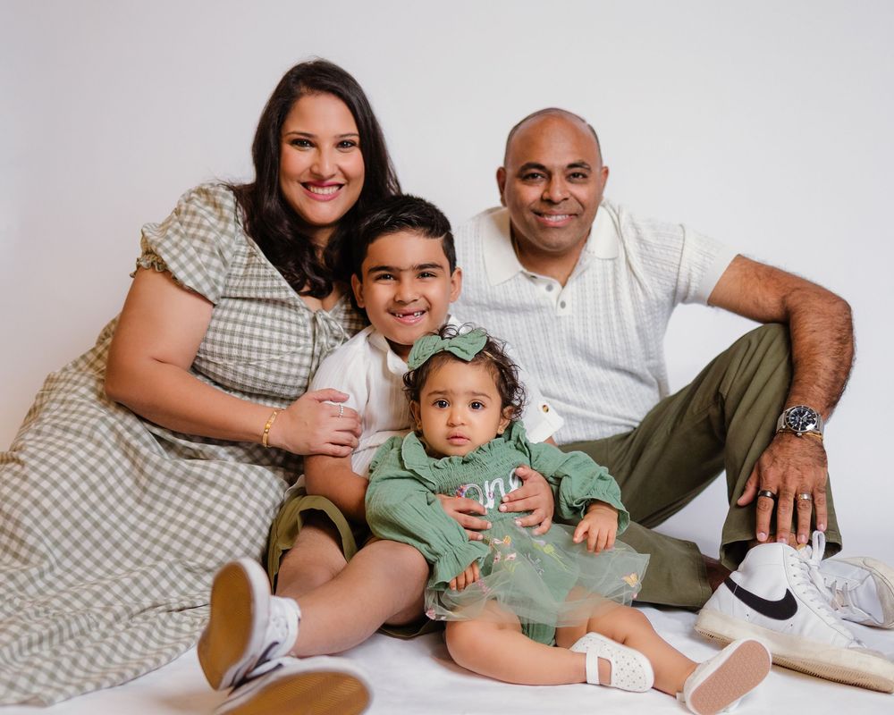 Family of four sitting casually together smiling in bright white studio setting.