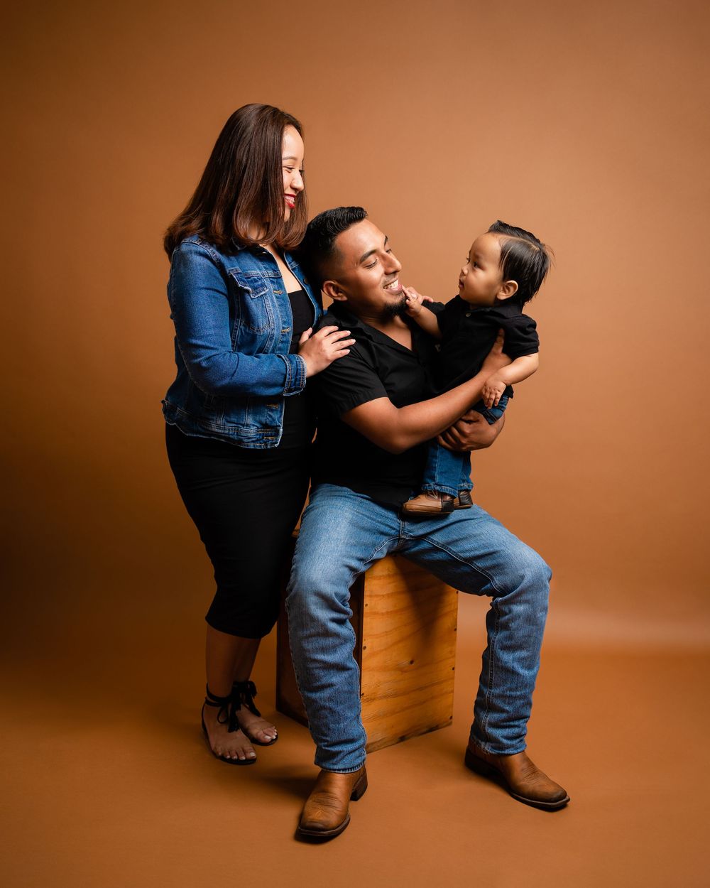 Family studio portrait with father seated holding baby while mother leans in on brown background.