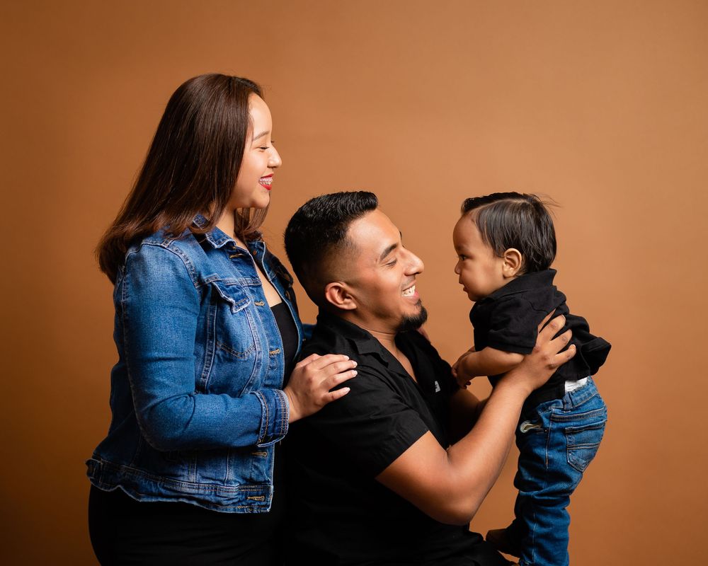 Studio family portrait of parents in denim outfits smiling at baby boy against warm brown background.