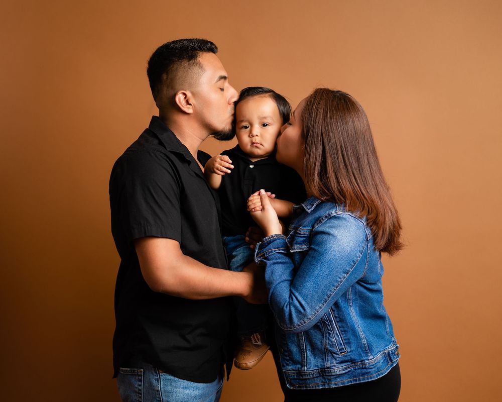 Loving parents in denim outfits each kissing their baby on cheeks in warm-toned studio portrait.