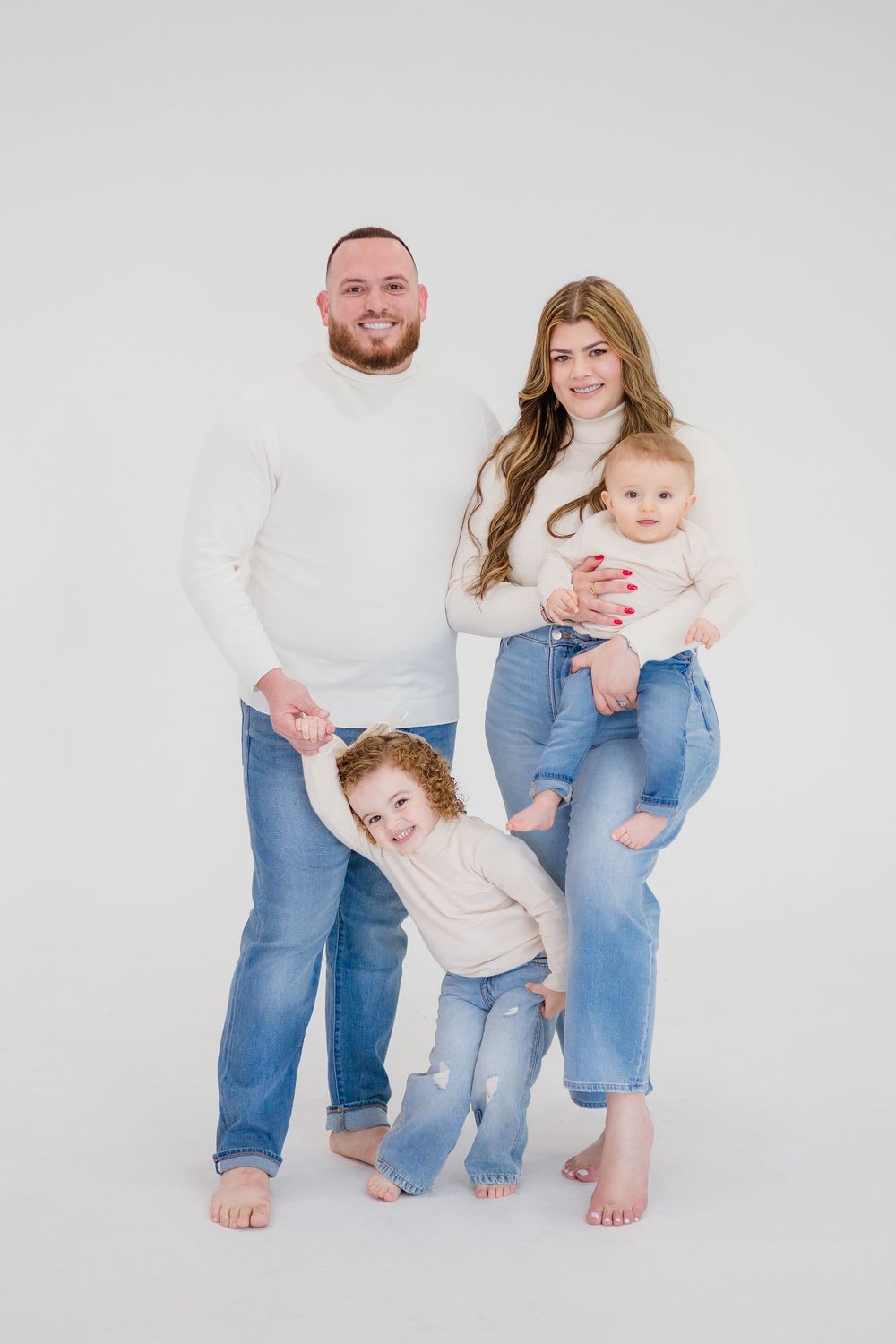 Family of four in matching white sweaters and jeans posing together on white studio background.