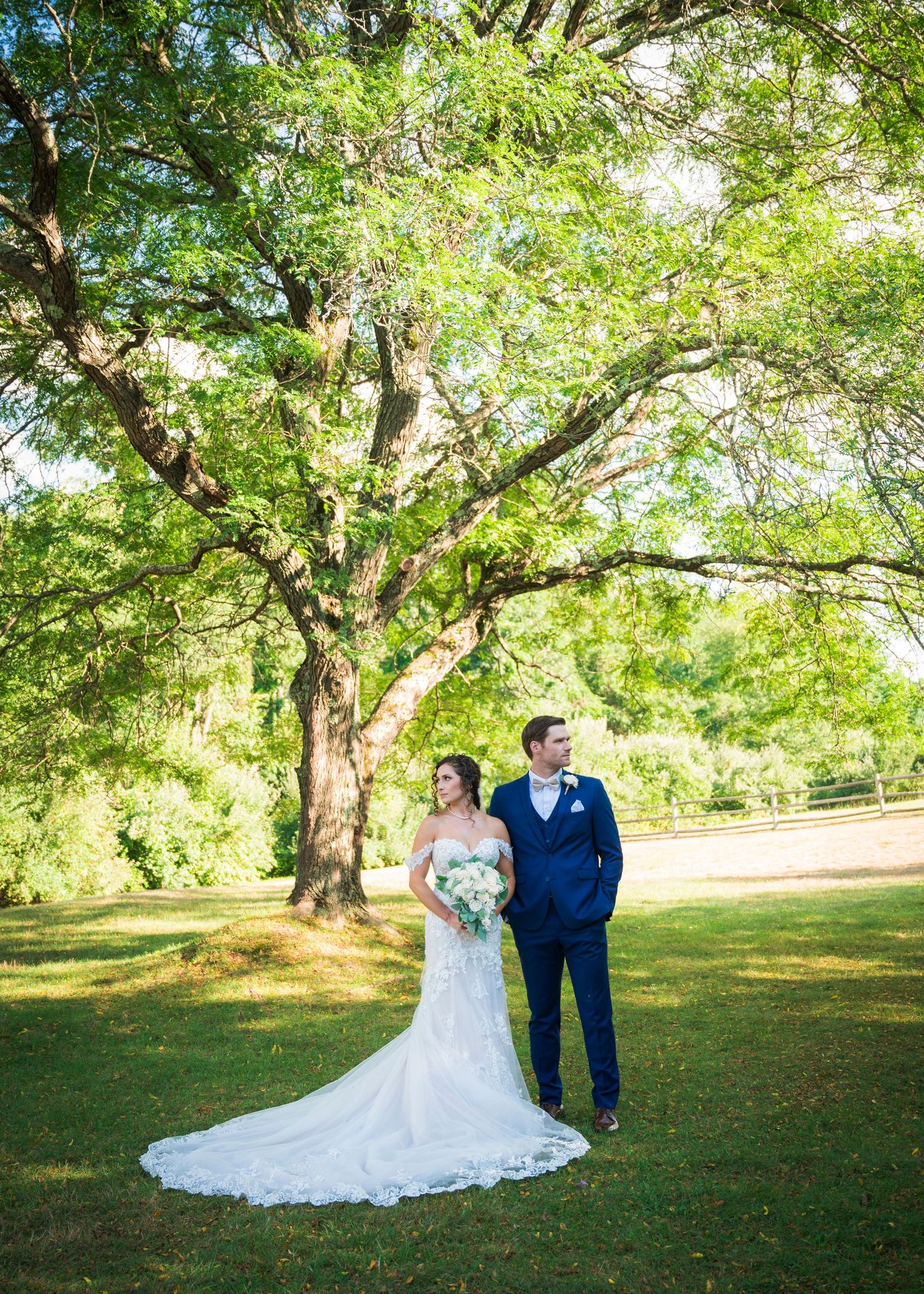 A couple shares a romantic moment under towering green trees in a serene park setting during their wedding celebration.