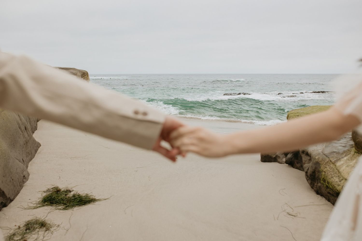 Two hands reach out to hold each other on a sandy beach with ocean waves in the background.