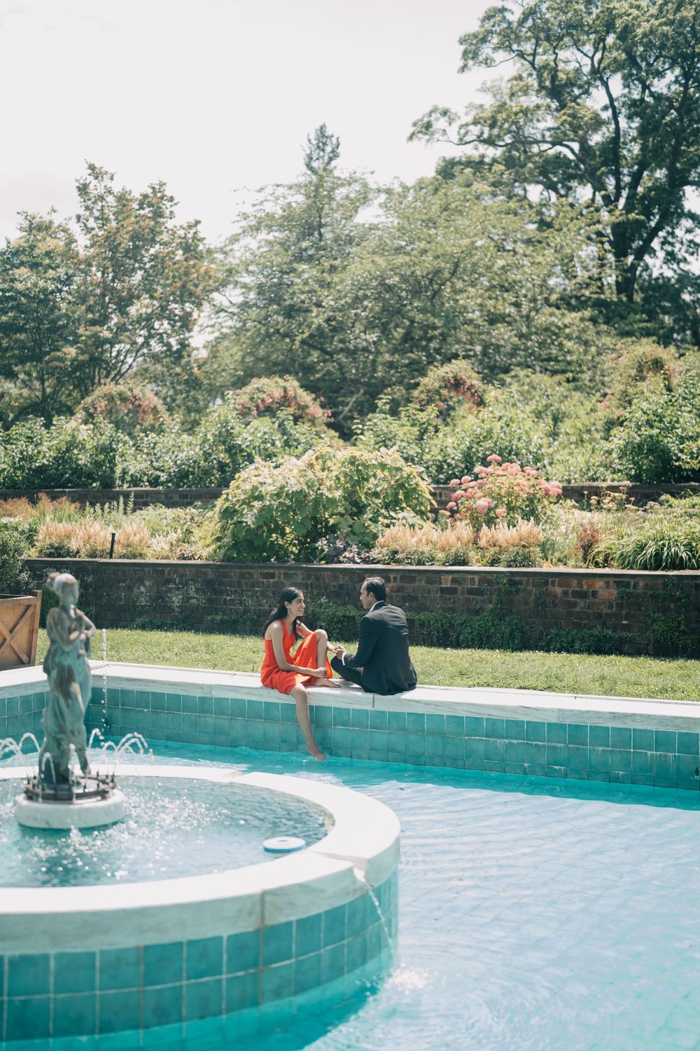 People enjoy a sunny day relaxing by a curved turquoise swimming pool surrounded by lush greenery and trees.