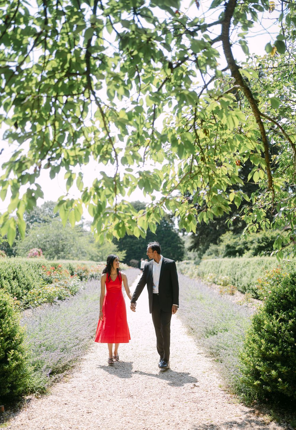 A couple walks hand in hand down a garden path surrounded by lush greenery and flowers on a sunny day.