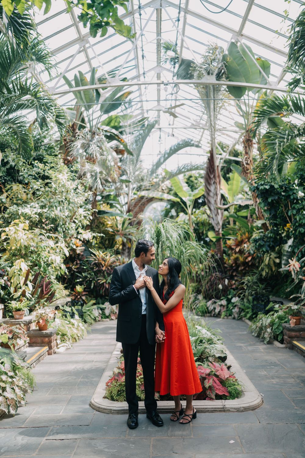 A couple shares romantic moments in a lush greenhouse conservatory filled with tropical plants and natural light.