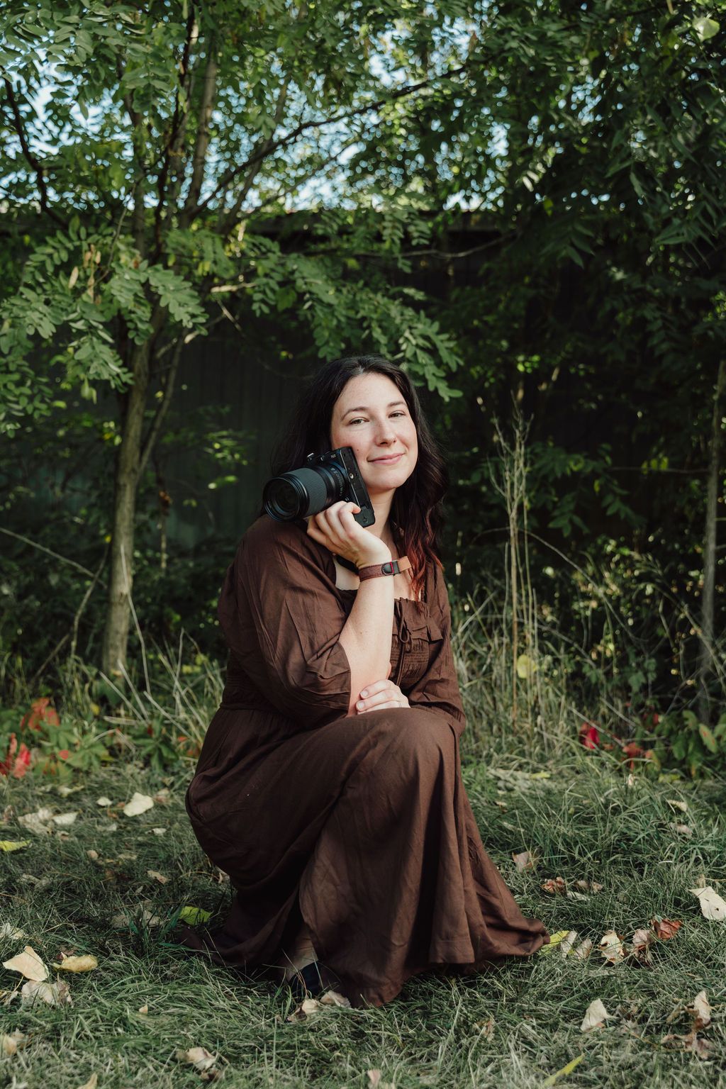 Someone in a brown dress sits thoughtfully in a garden setting surrounded by greenery and fallen leaves.