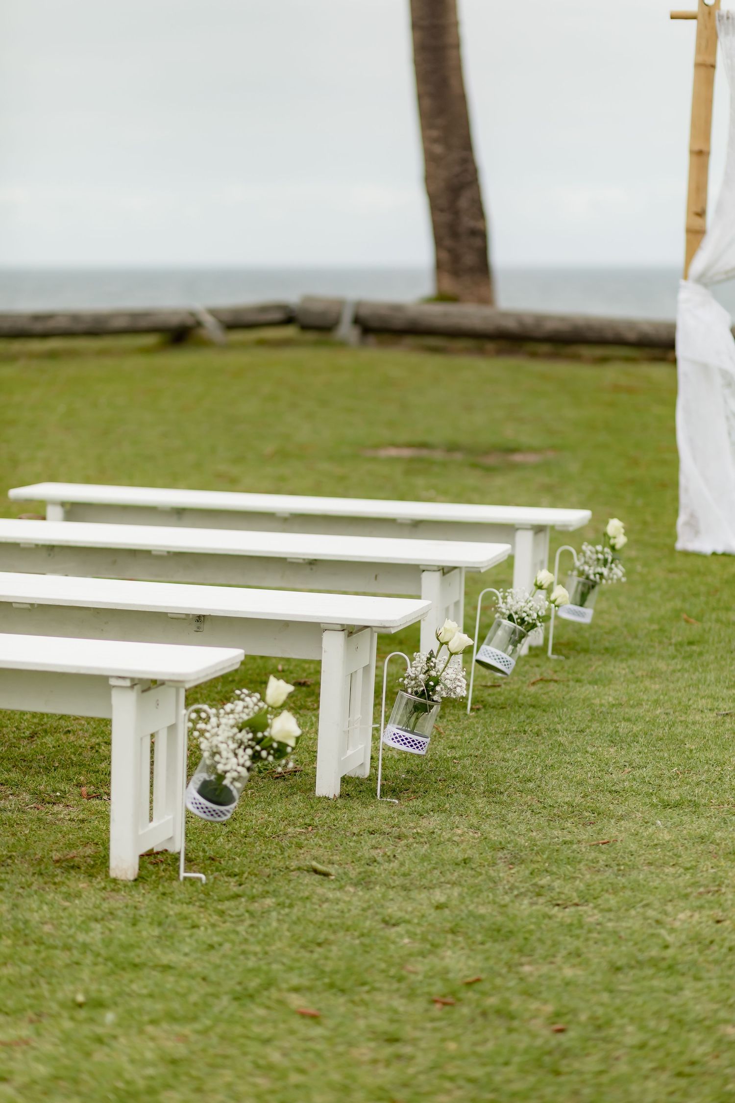 White wooden benches decorated with small floral arrangements line a grassy outdoor wedding ceremony space.