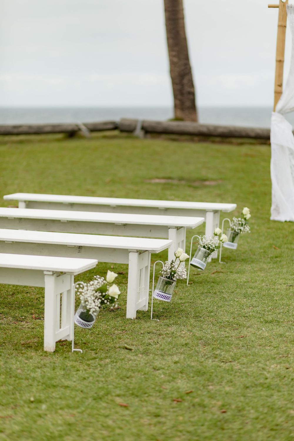 White wooden benches decorated with small floral arrangements line a grassy outdoor wedding ceremony space.