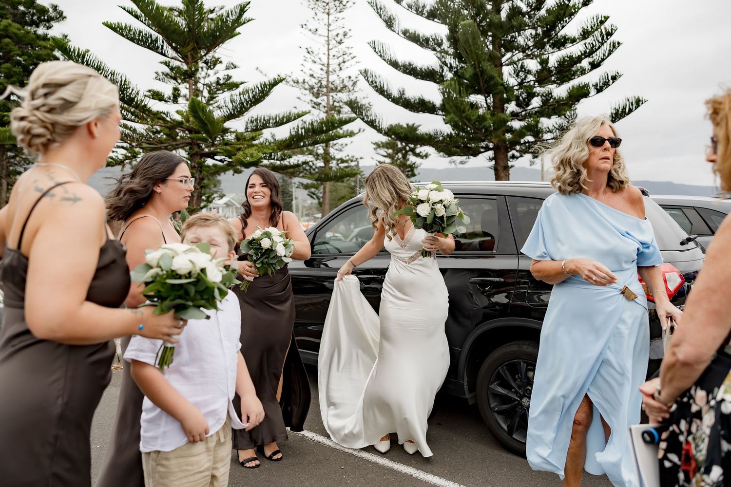 Wedding party gathers in a parking lot with pine trees and cars in the background.