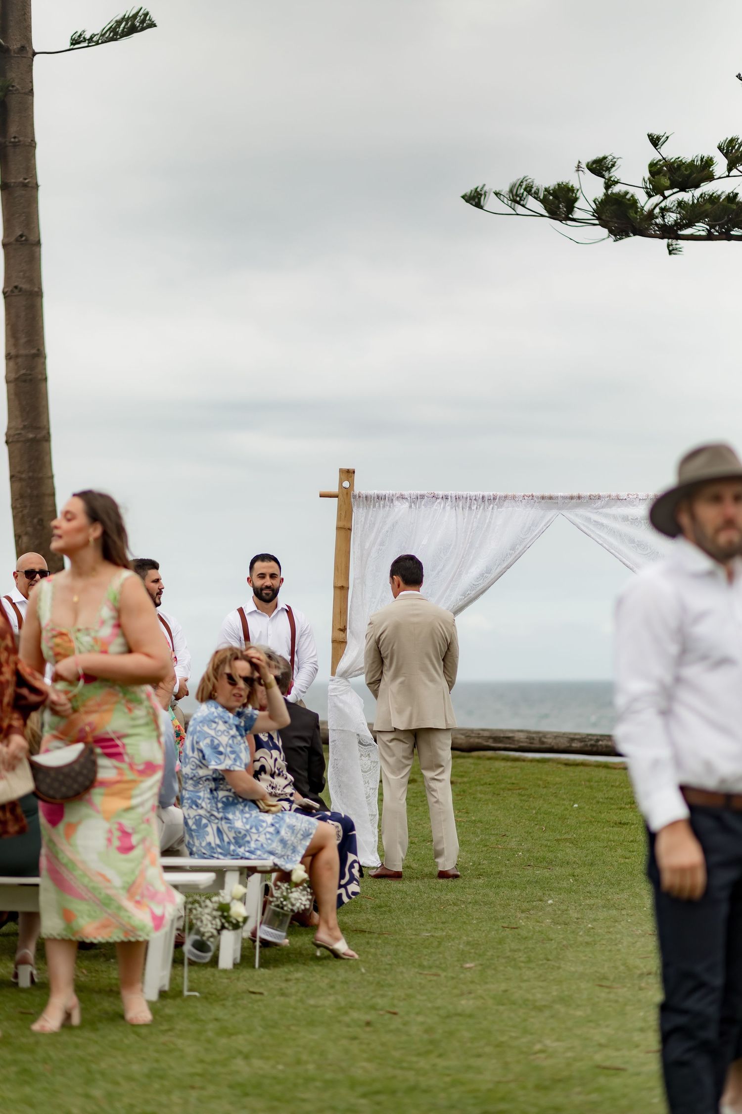 Guests gather at an outdoor wedding ceremony setup with white fabric arch overlooking the ocean.