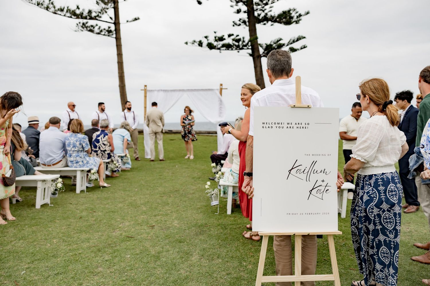 Outdoor wedding ceremony setup with white benches and decorative signage against an ocean backdrop.