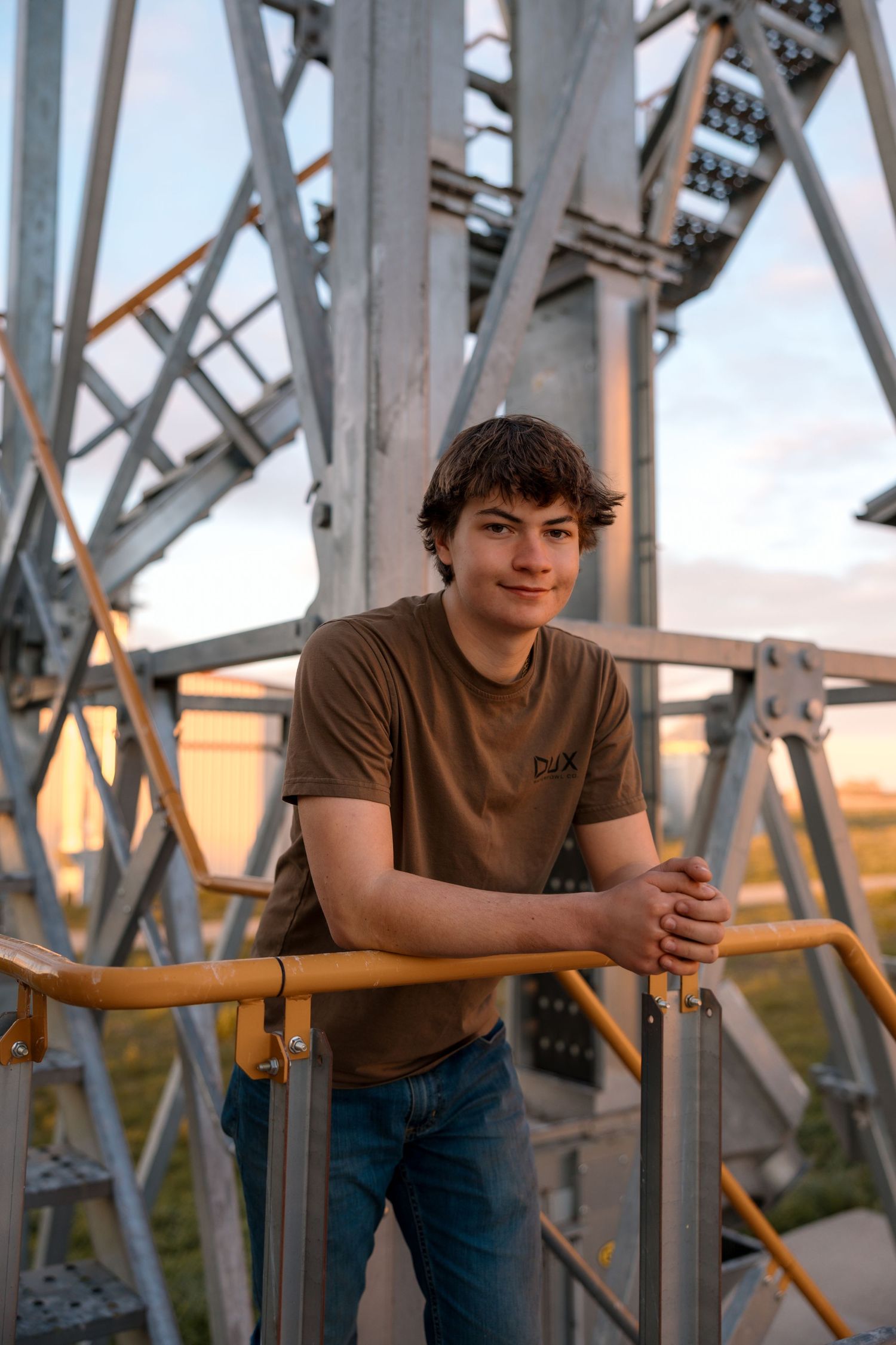 Portrait of someone in a brown t-shirt leaning on railing with industrial metal bridge structure in background at sunset.