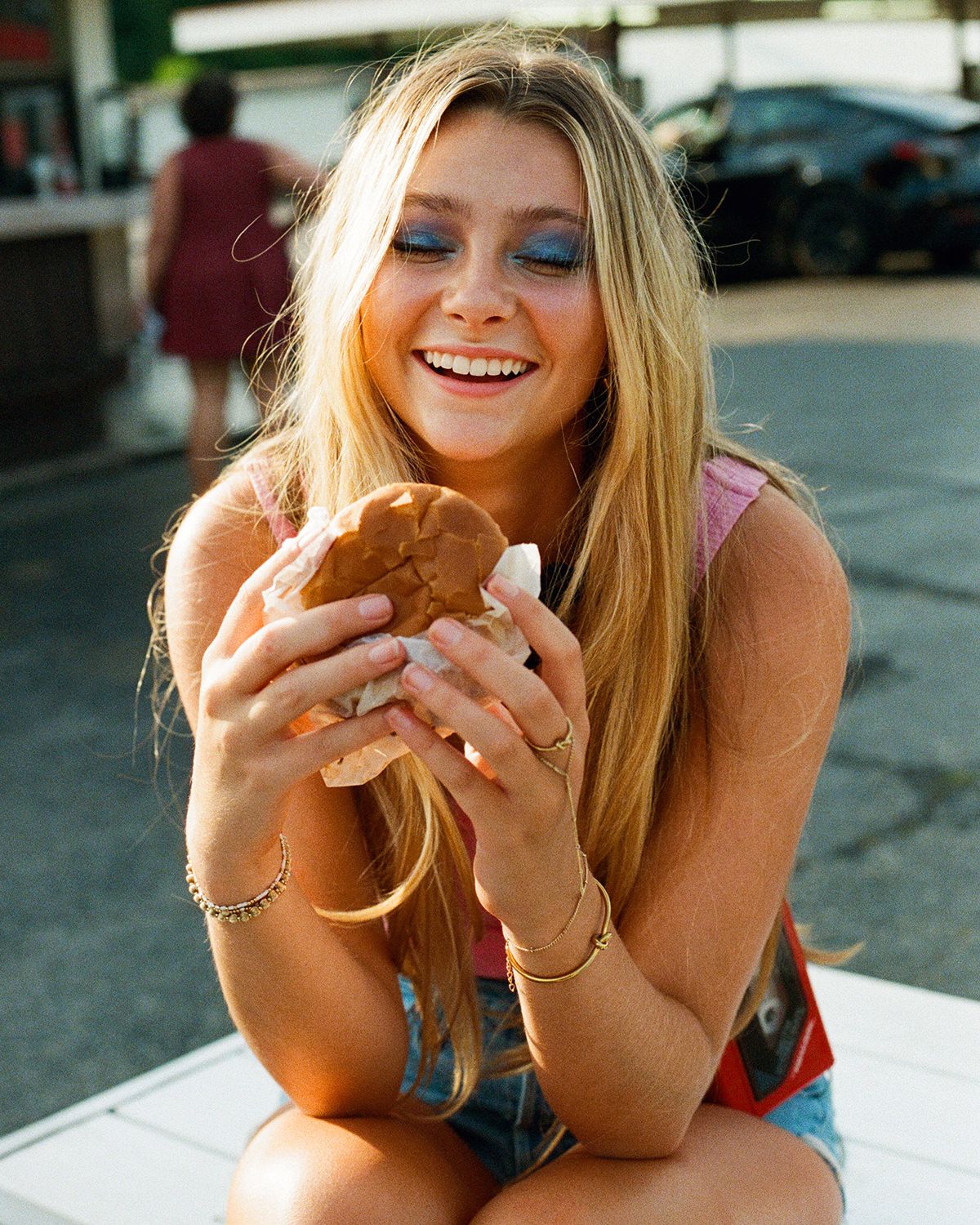 A woman with blonde hair smiles while enjoying a cheeseburger outdoors.