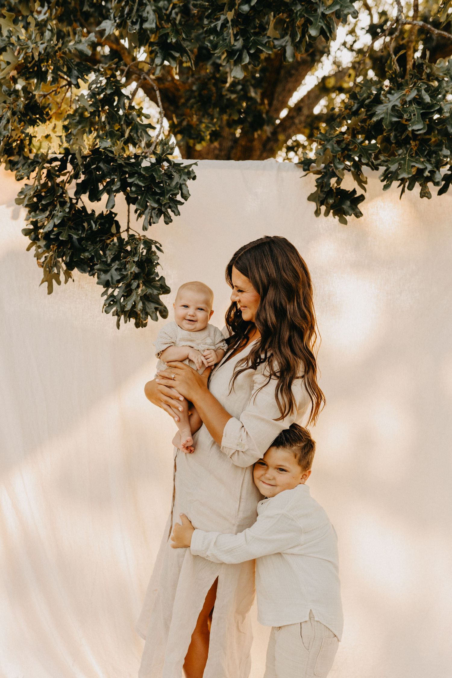 A mother holds her baby while her young child embraces her, standing beneath a lush tree outdoors.