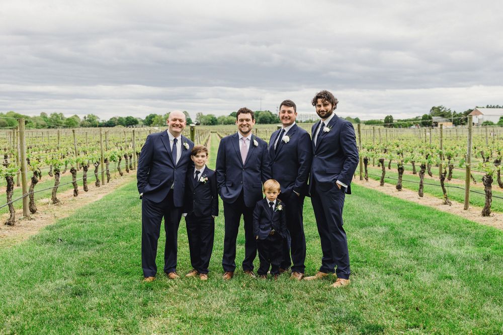 Wedding party poses together in navy suits between rows of grapevines at a scenic vineyard on a sunny day.