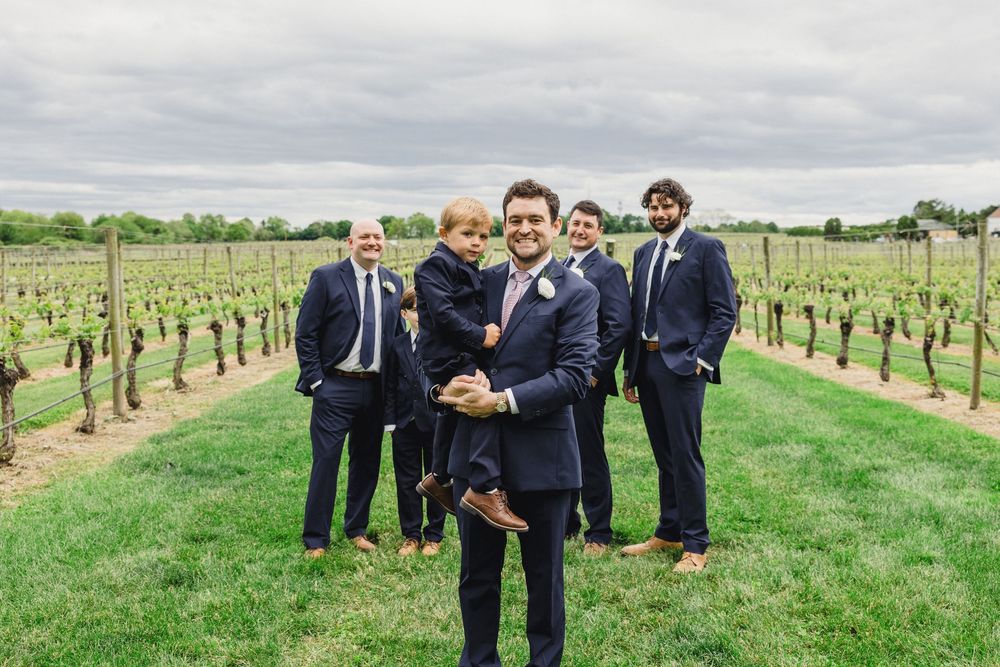 Groomsmen in navy suits pose together in a vineyard on a cloudy day.