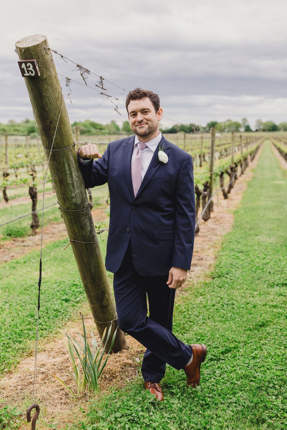 Groom in navy suit poses playfully near vineyard posts.