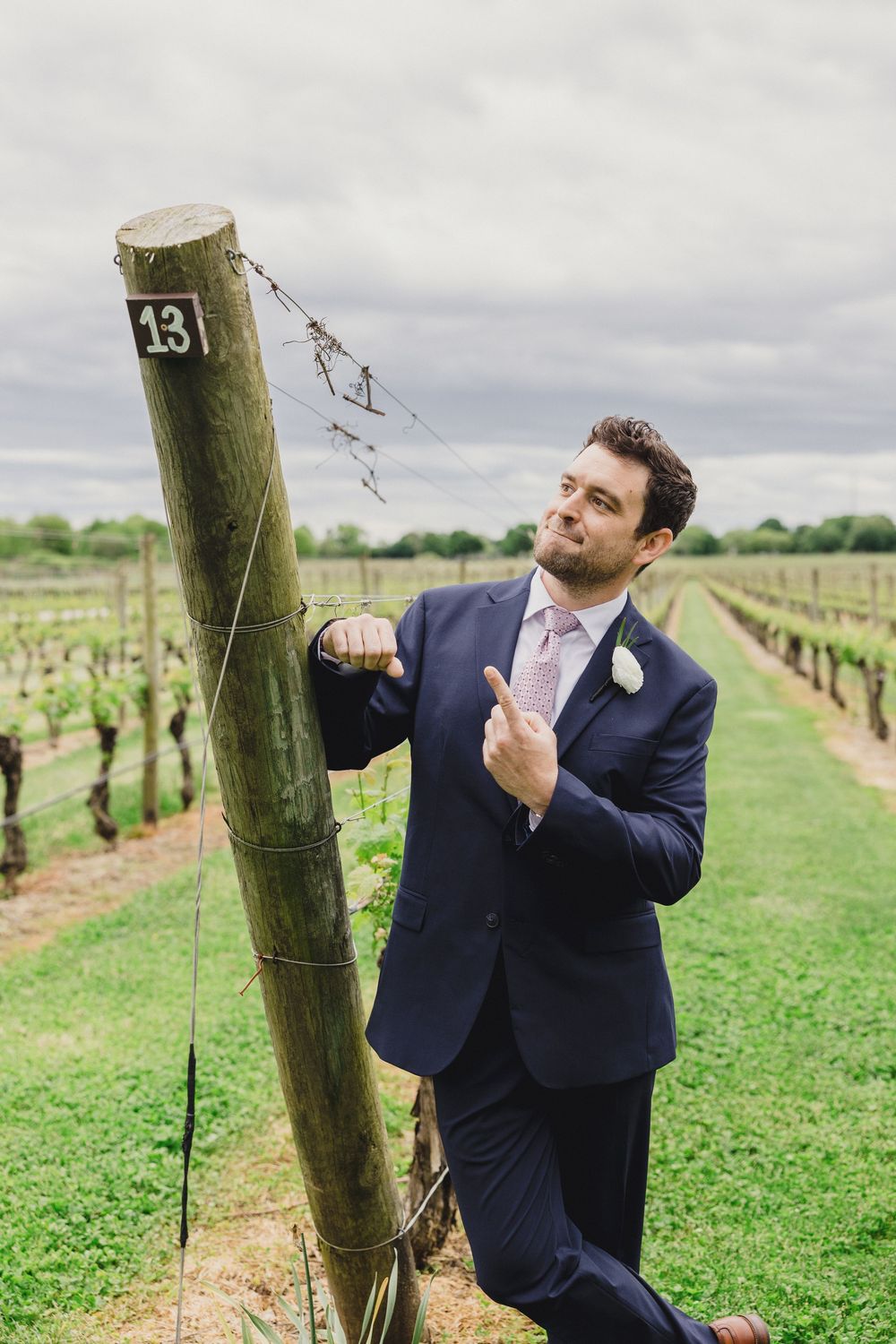 Groom in dark suit making funny poses between vineyard rows.