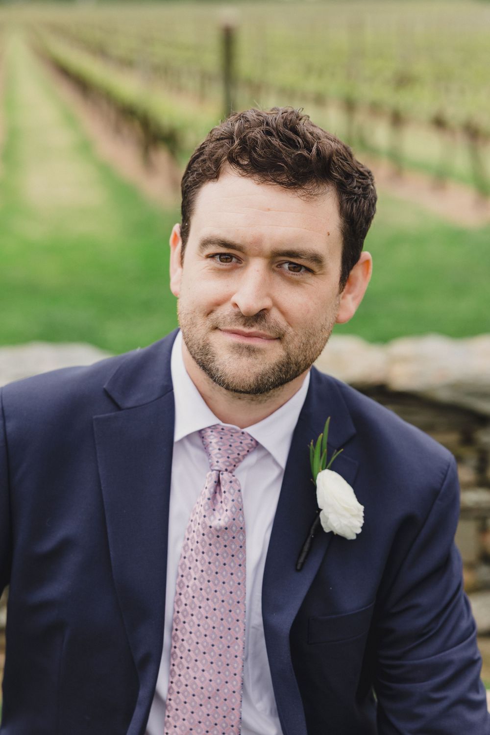 A groom in a navy suit with pink patterned tie and white boutonniere smiles at the camera in a vineyard setting.