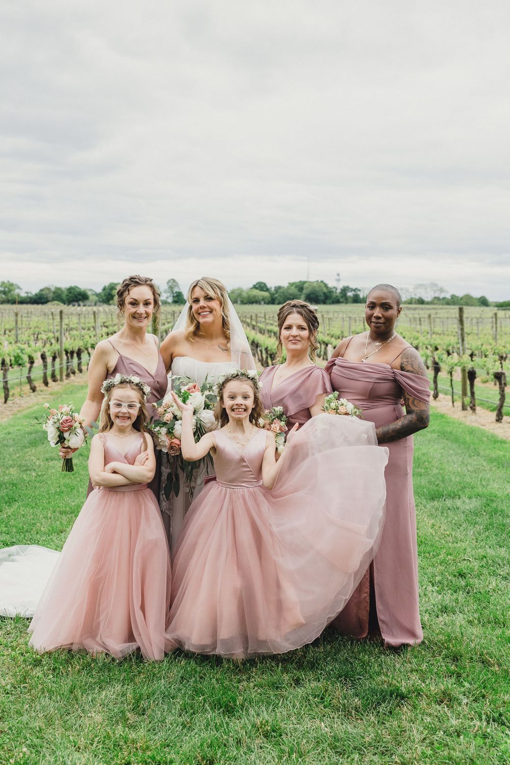 Bridesmaids in dusty rose dresses pose together in a vineyard setting.