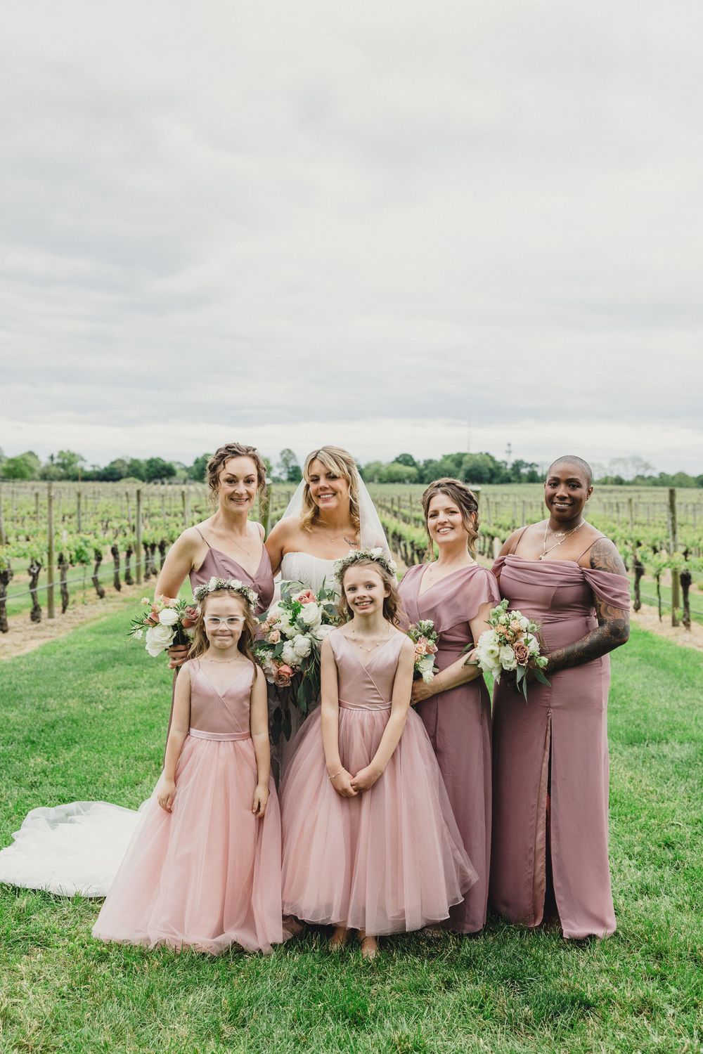 Wedding party in pink dresses stands together against a backdrop of vineyard rows.