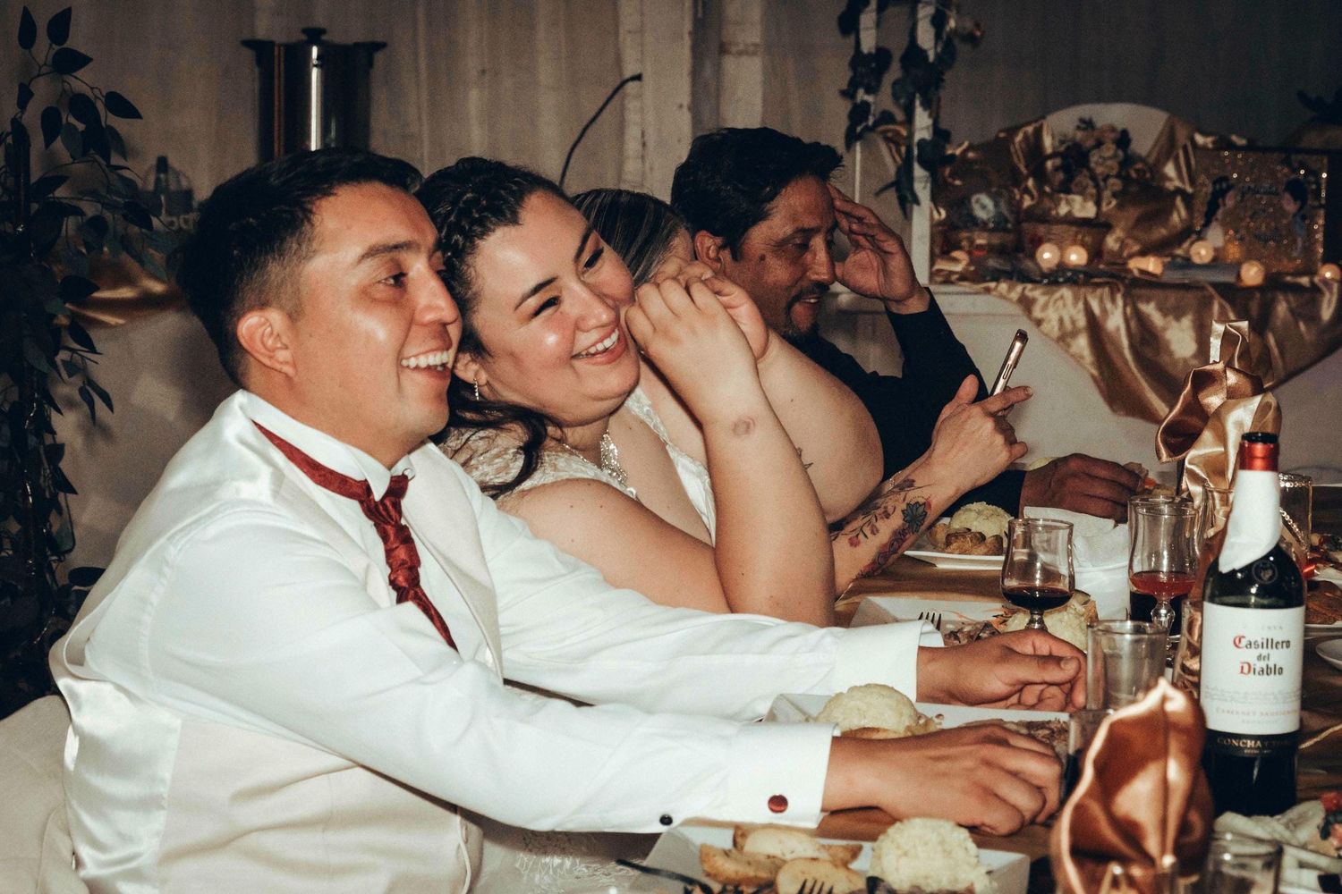 Invitados en recepción de matrimonio sentados en largas mesas disfrutando la cena en ambiente tenue.