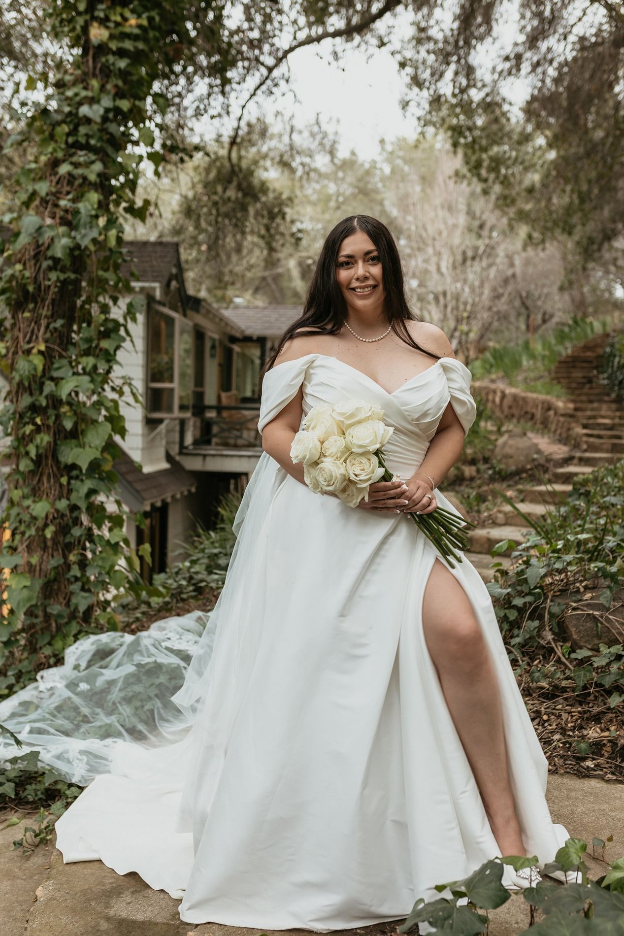 A white wedding dress with an off-shoulder neckline flows elegantly in a garden setting with stone steps and greenery.