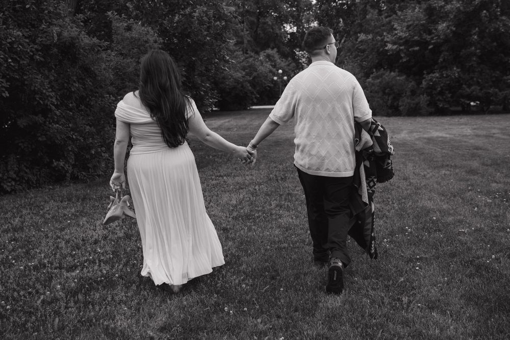 A couple holding hands walks away from the camera across a grassy field in black and white photography.