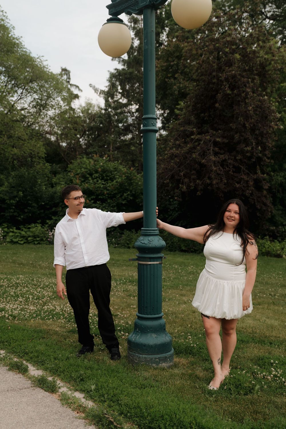 A couple poses playfully by a vintage-style green lamp post in a park setting with grass and trees.