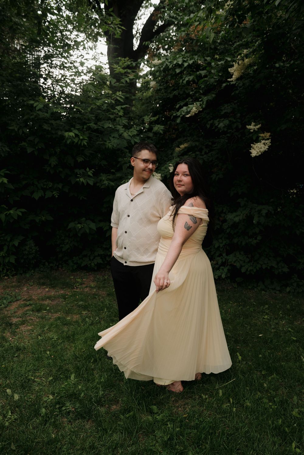 A couple in light-colored outfits twirls and dances in a green outdoor setting.