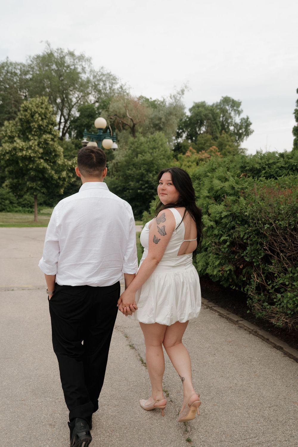 A couple in formal attire walking together on a garden path, with the woman in a white dress looking back.