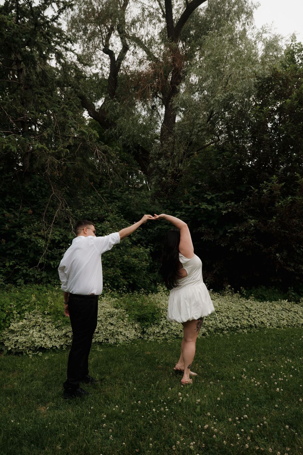 A couple dances playfully in a grassy field surrounded by trees on a summer day.