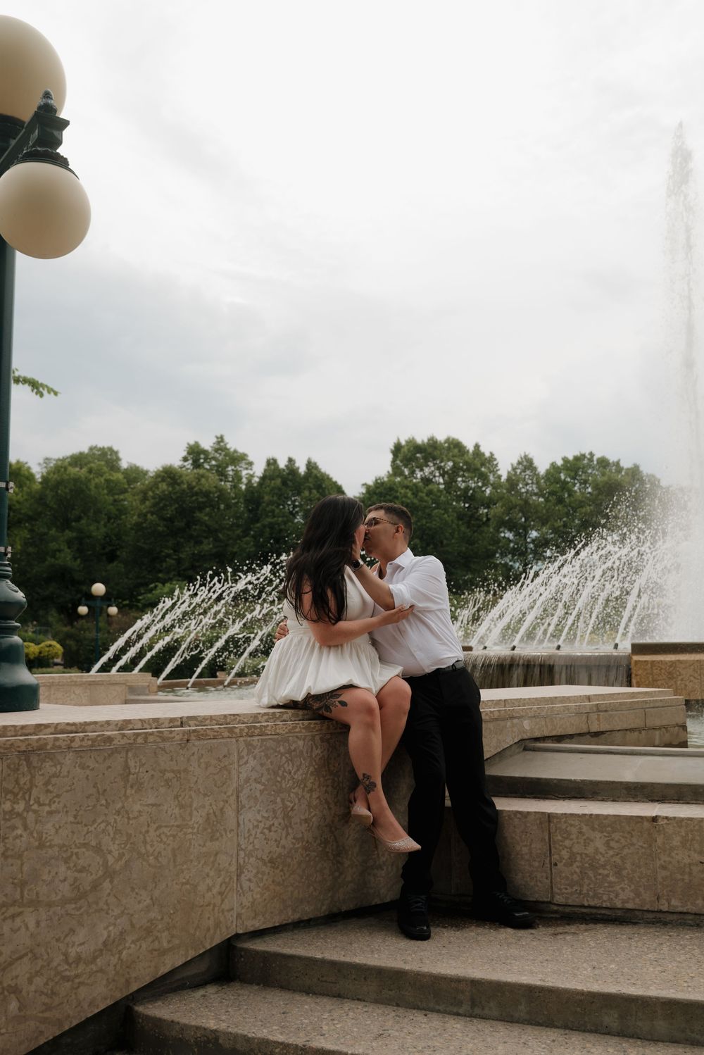 A romantic moment shared between a couple sitting by a fountain with water jets spraying in the background.