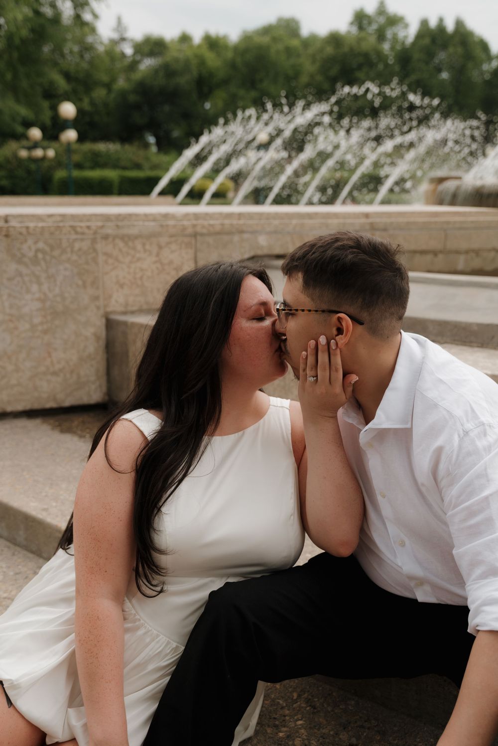 A romantic couple shares an intimate moment sitting by a fountain in a park setting.