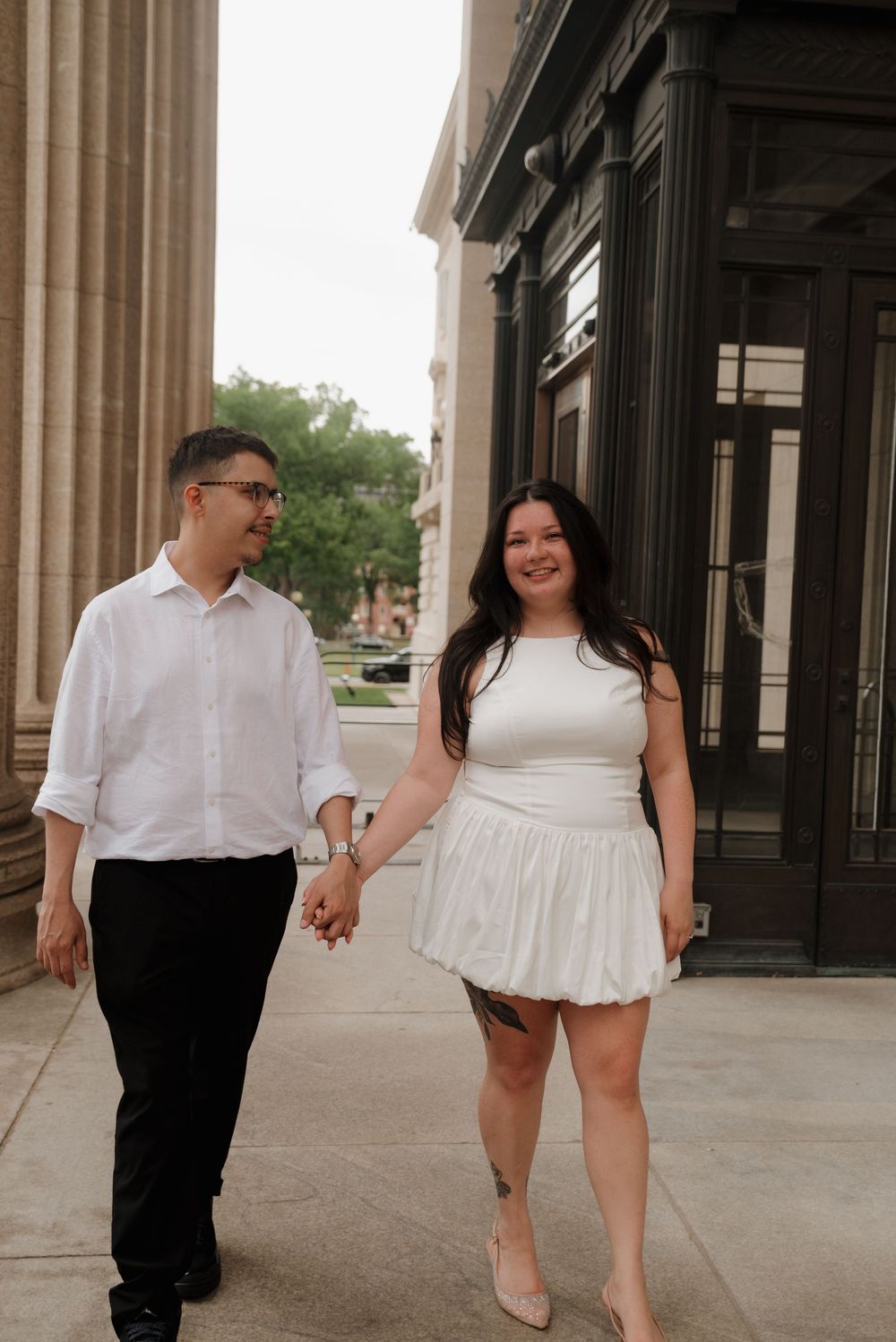A couple in white and black attire hold hands while walking outside a building with classical architecture.