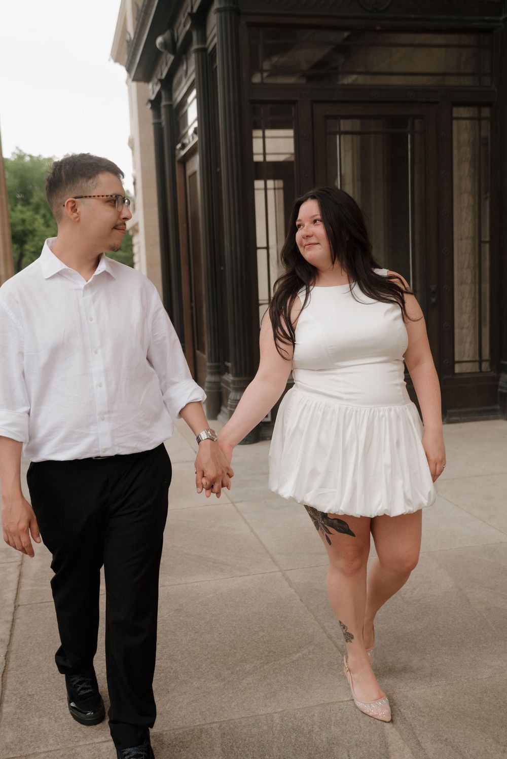 A couple holding hands while walking together outside a building, with one person in white and one in dark attire.