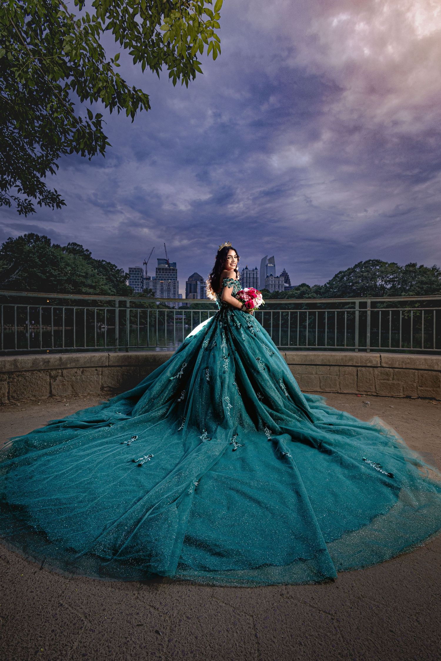 A dramatic teal ballgown with a sweeping train flows across a stone terrace with a cityscape backdrop at dusk.