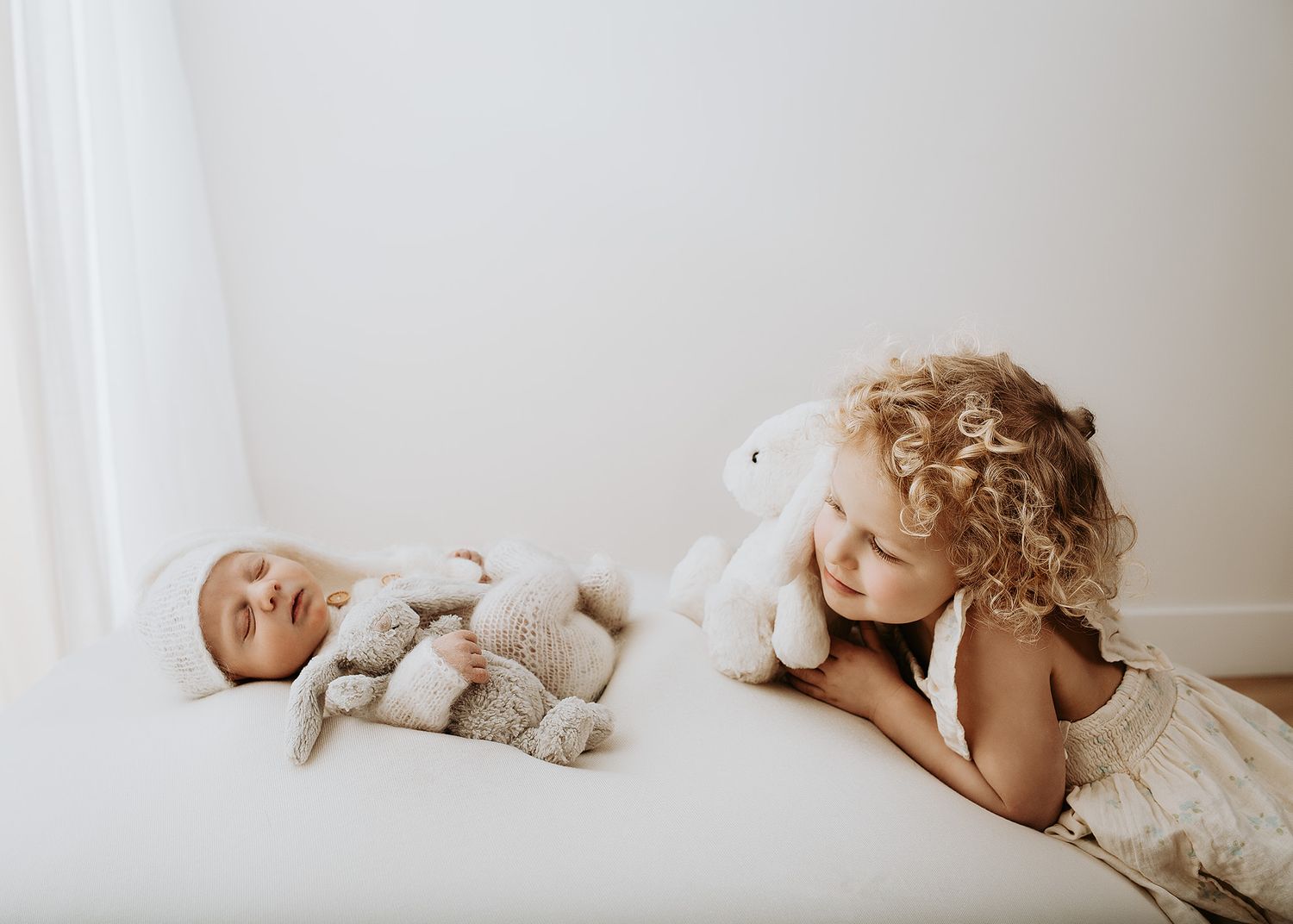 Curly-haired toddler gently looking at her newborn baby sibling lying on a cream bed in a light and airy studio newborn session.