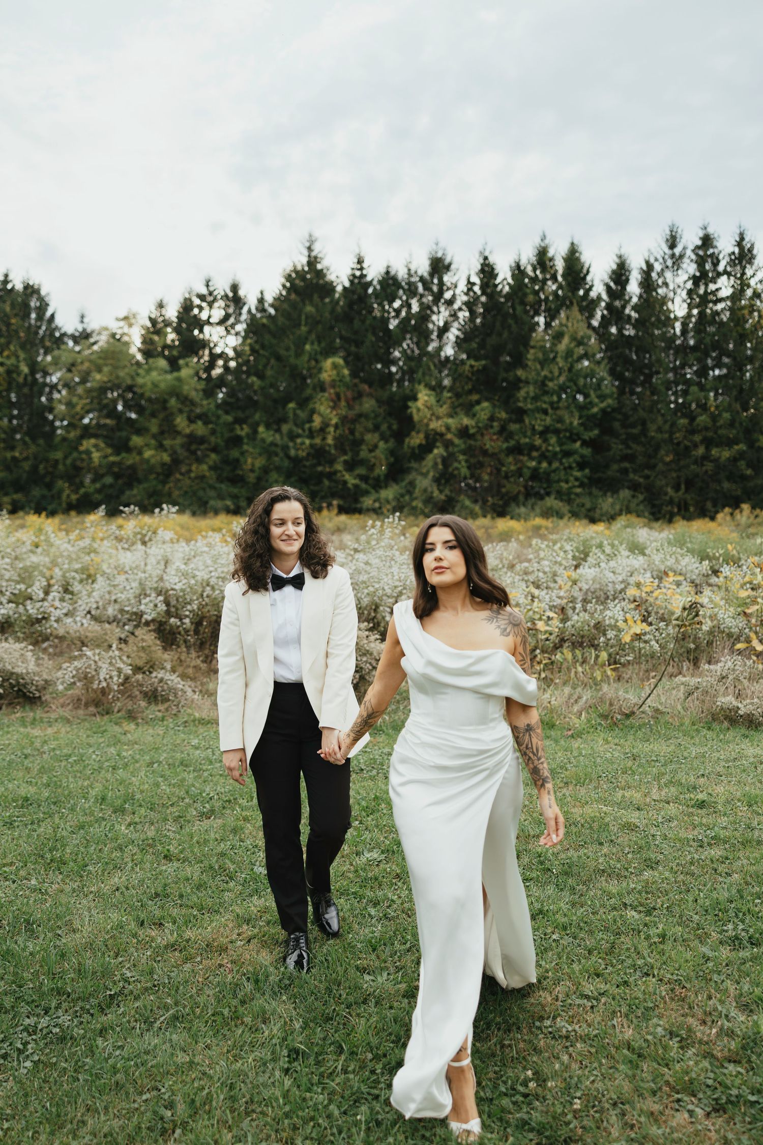 A couple in wedding attire walks and dances playfully together in a meadow with evergreen trees in the background.