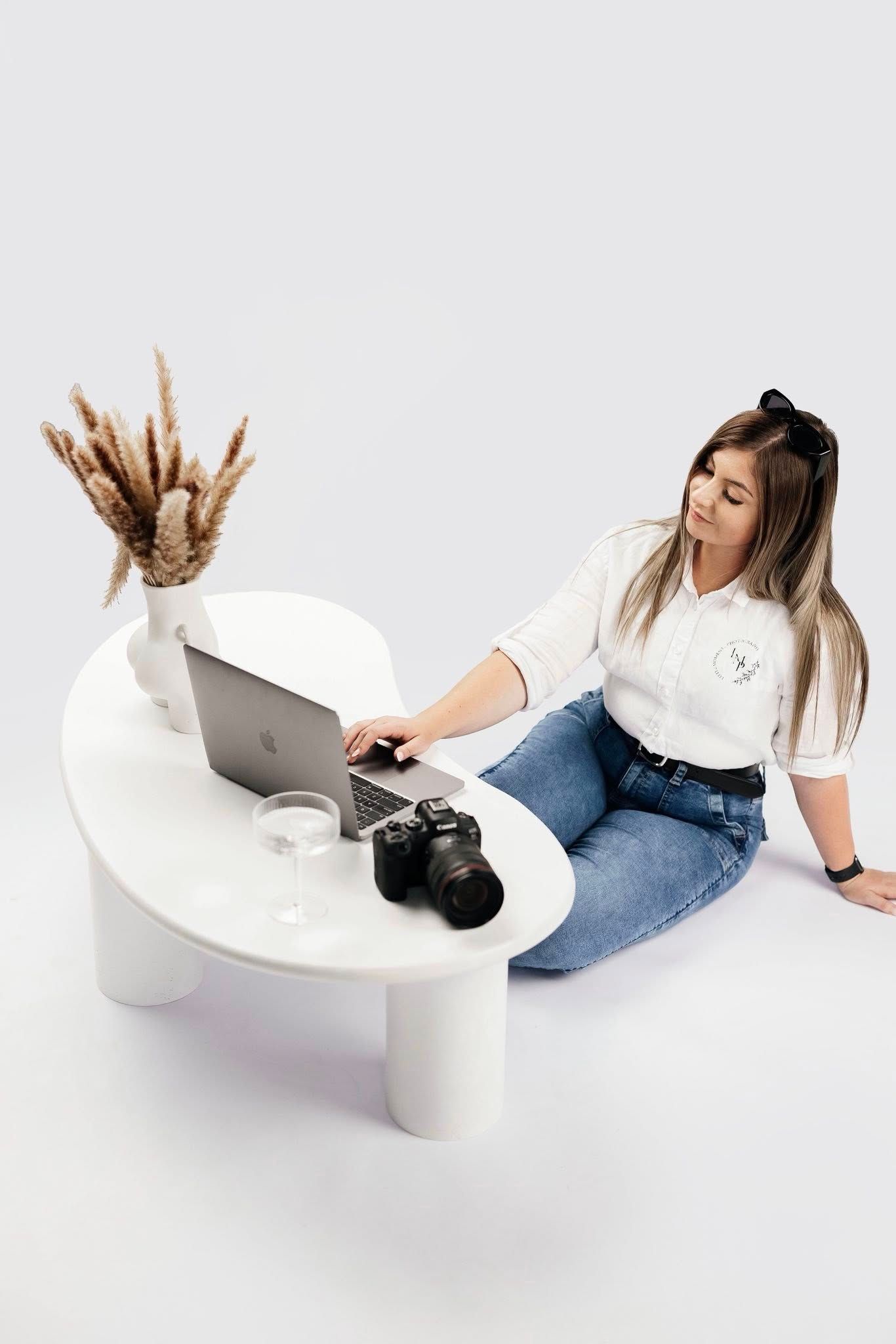 Female photographer working on laptop beside DSLR camera at white round studio table.