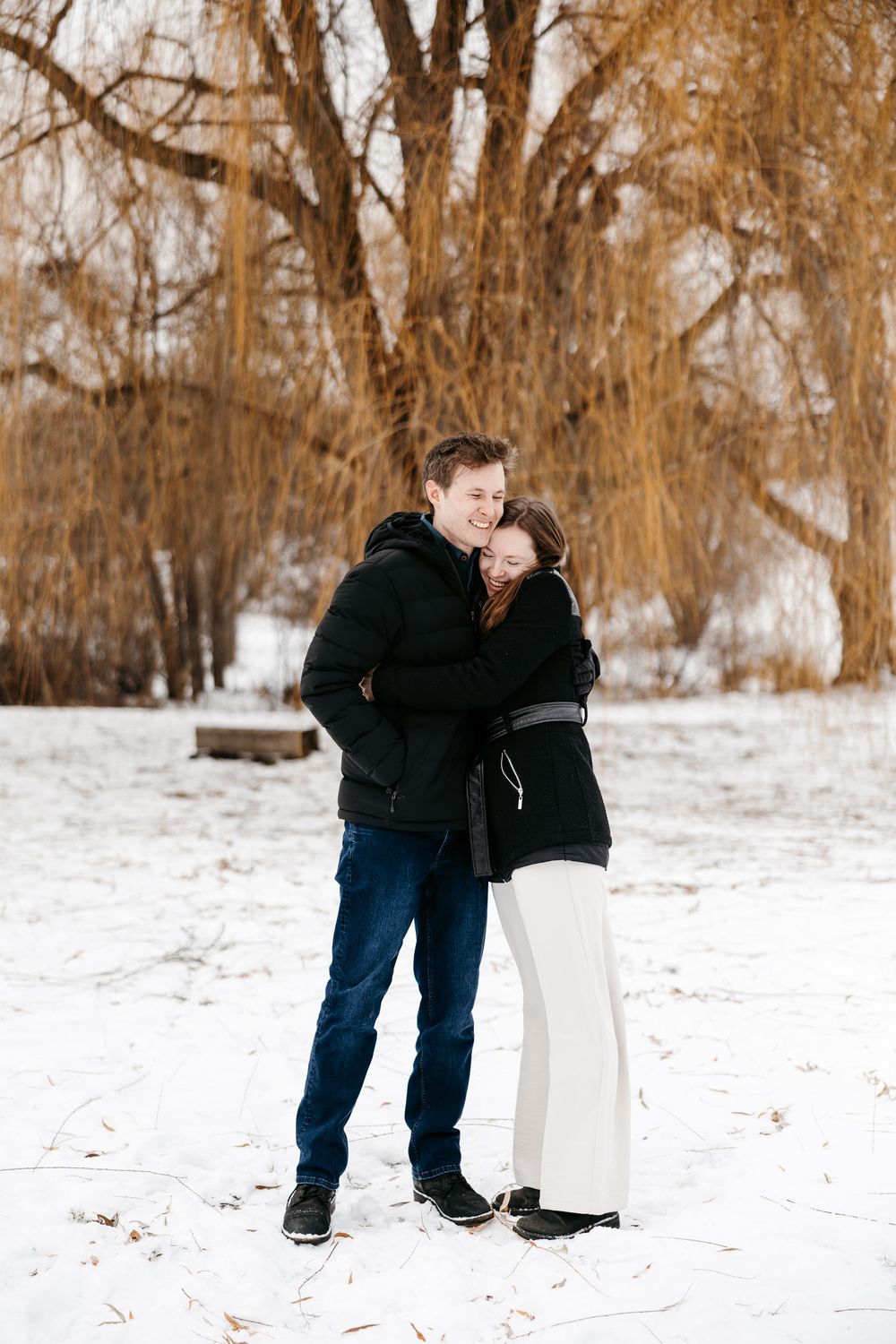 A couple embraces while standing in a snowy winter landscape with bare trees in the background.