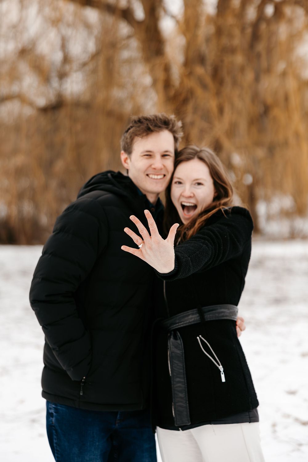 Two people share joyful moments together while standing in snow covered landscape with brown trees.