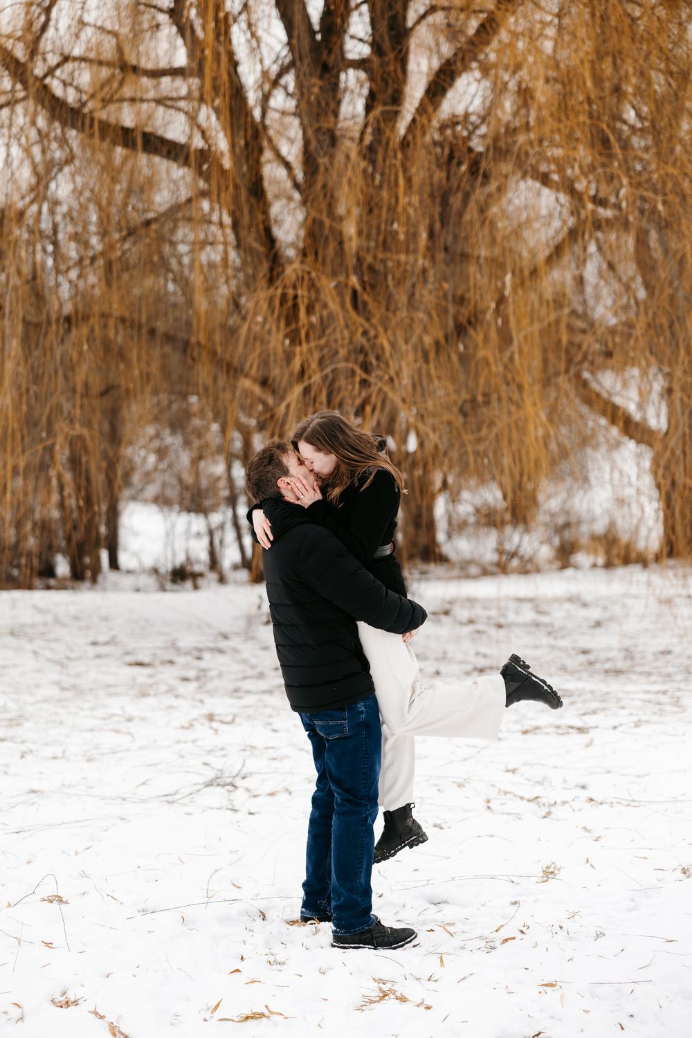 A romantic winter scene showing a couple hugging in front of snow-covered trees with warm brown foliage.