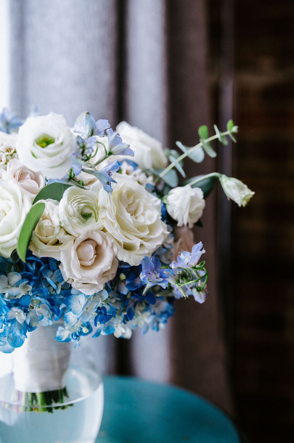 Elegant wedding bouquet featuring white roses and blue delphinium flowers in a glass vase against a dark background.