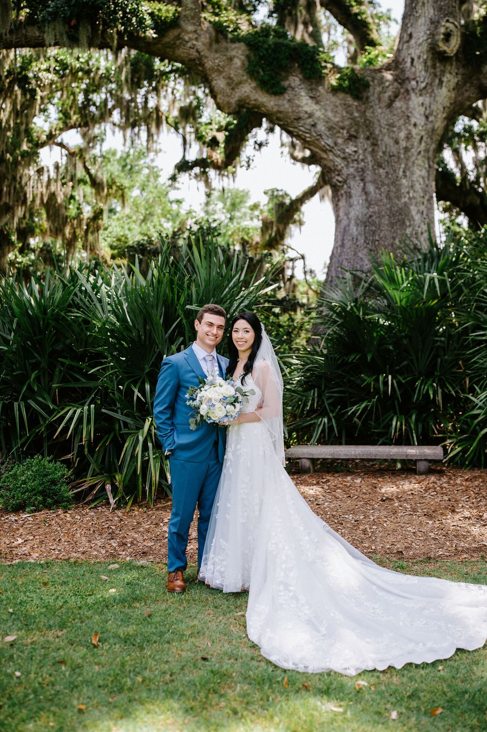 A couple shares a romantic moment in a lush garden setting with Spanish moss draped trees and tropical foliage.