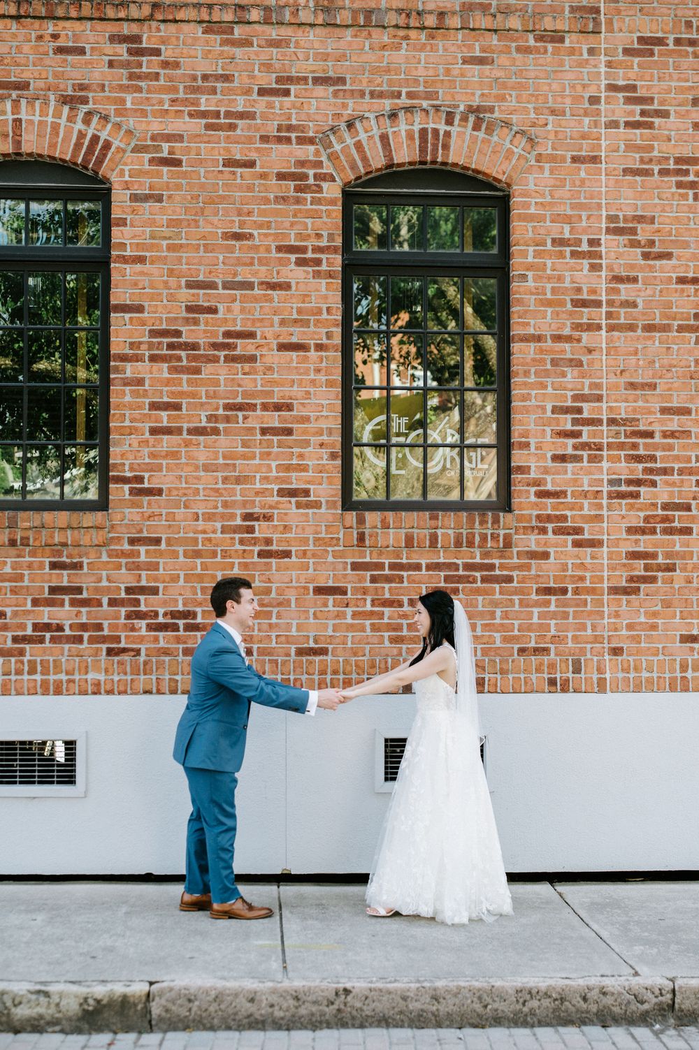 A couple in wedding attire holds hands against a rustic brick wall with arched windows.