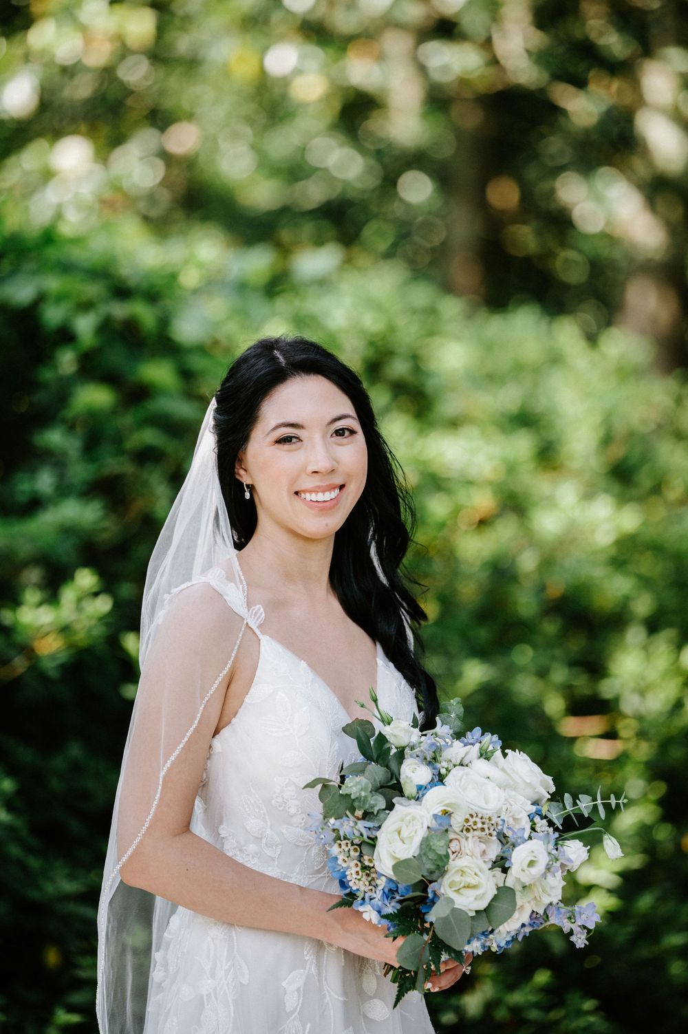 A bride in a white lace dress holds a delicate bouquet of white and blue flowers against a natural outdoor backdrop.