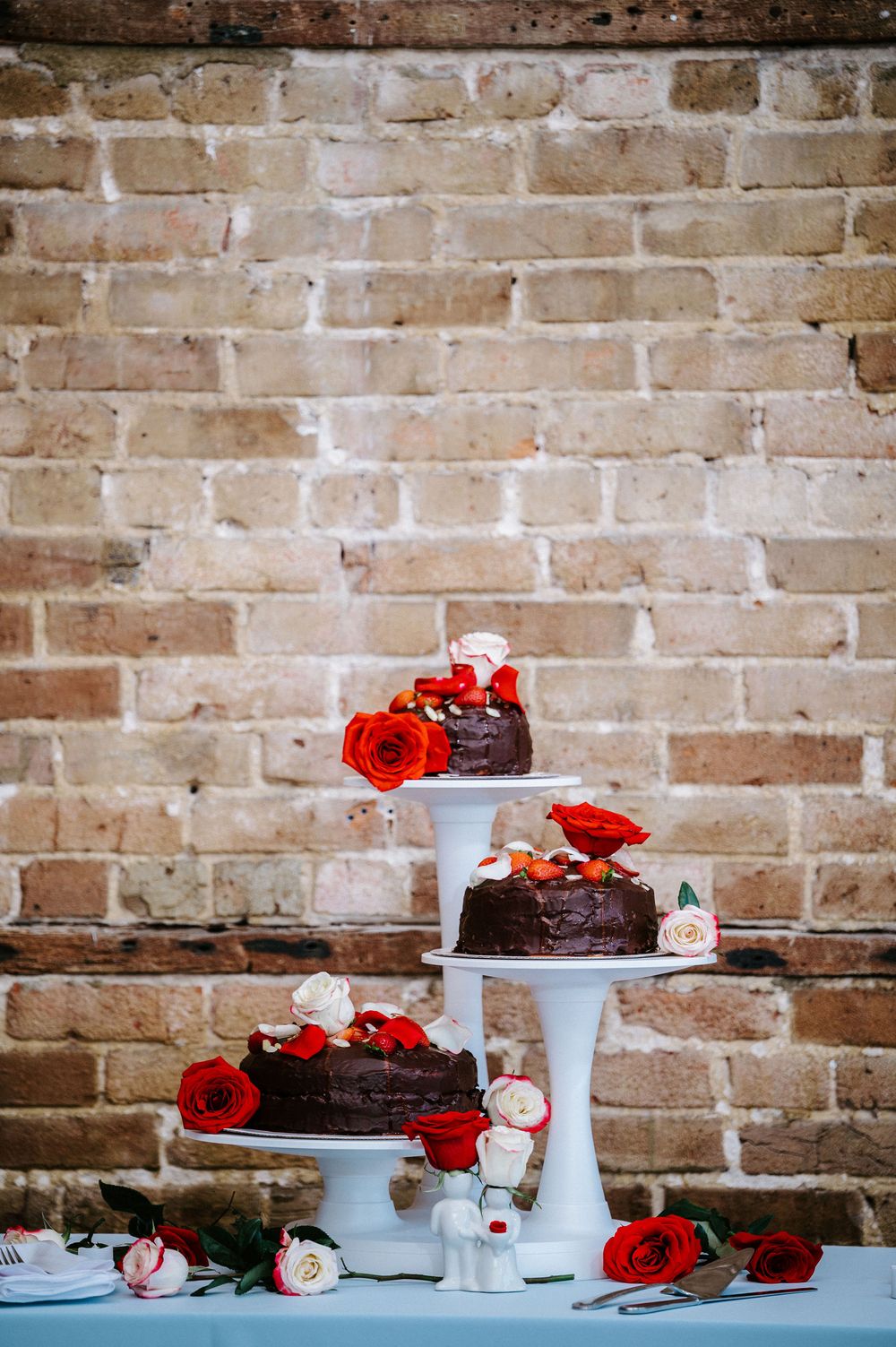 Chocolate cakes decorated with red flowers displayed on blue cake stands against a rustic brick wall.
