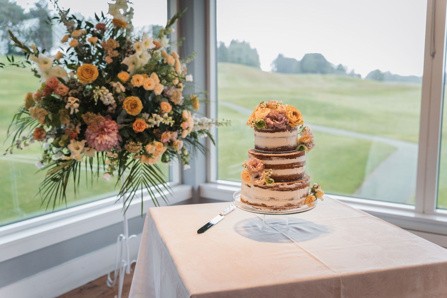 Rustic naked wedding cake decorated with fresh flowers and greenery sits by a window overlooking rolling hills.